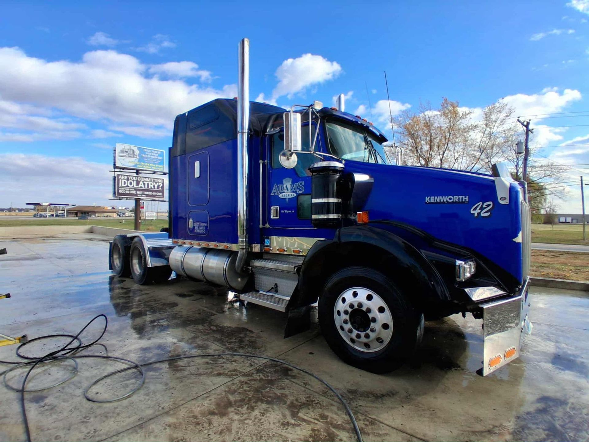 Blue semi-truck parked on a wet surface under a bright sky.