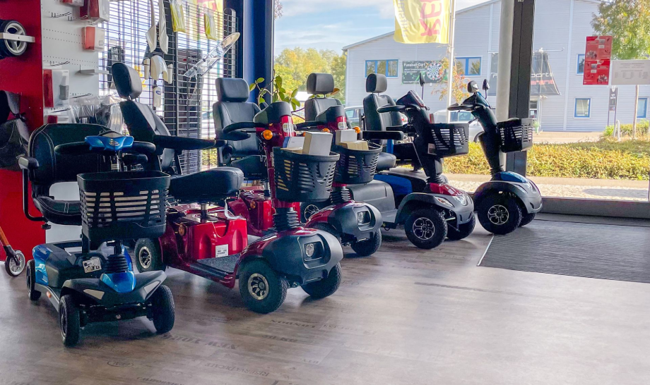 Bei Reha-Trend in Speyer stehen mehrere Elektromobile in verschiedenen Farben in einer Reihe, nahe einem Schaufenster.