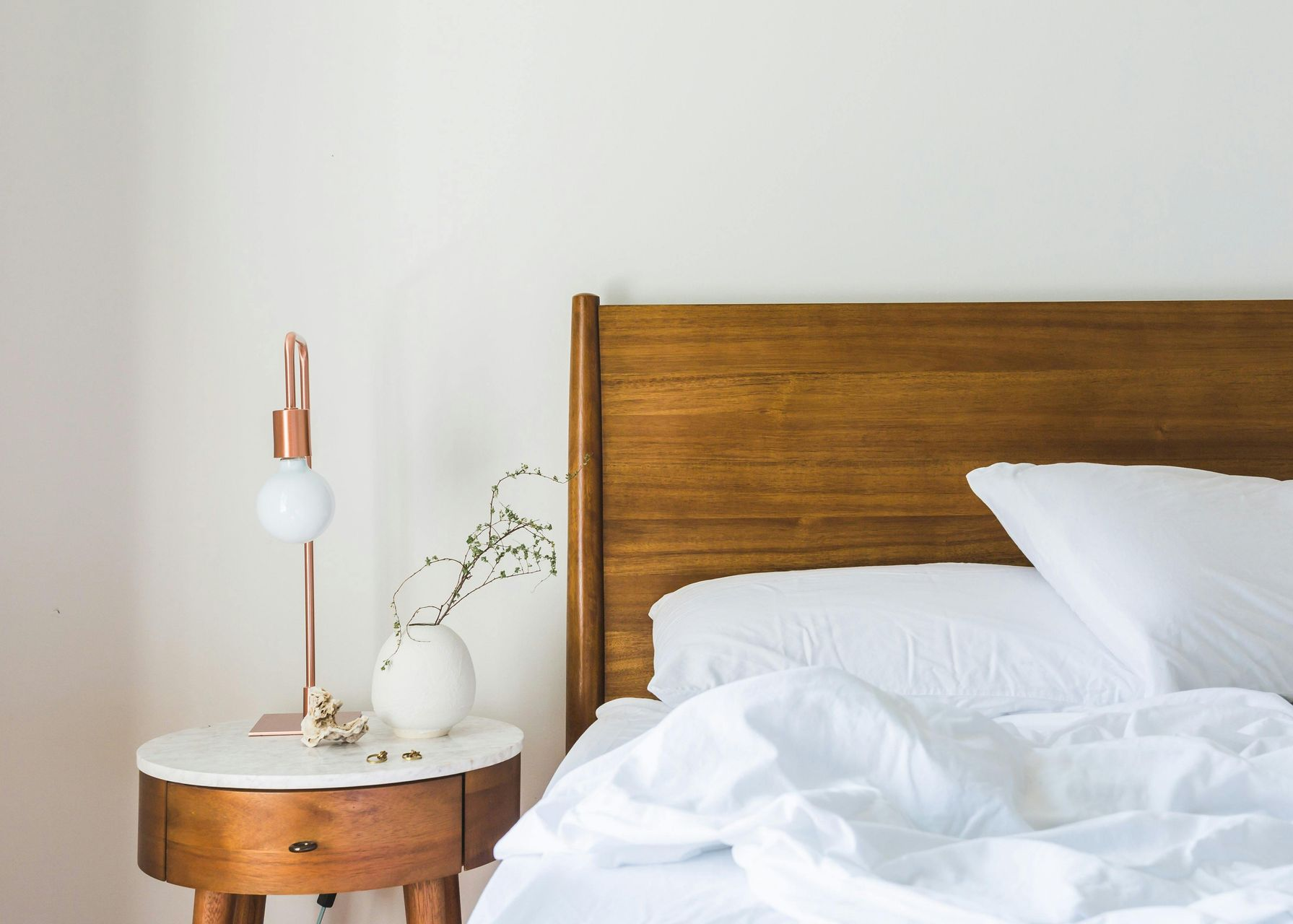 Wooden bed with white bedding, nightstand with lamp and vase.