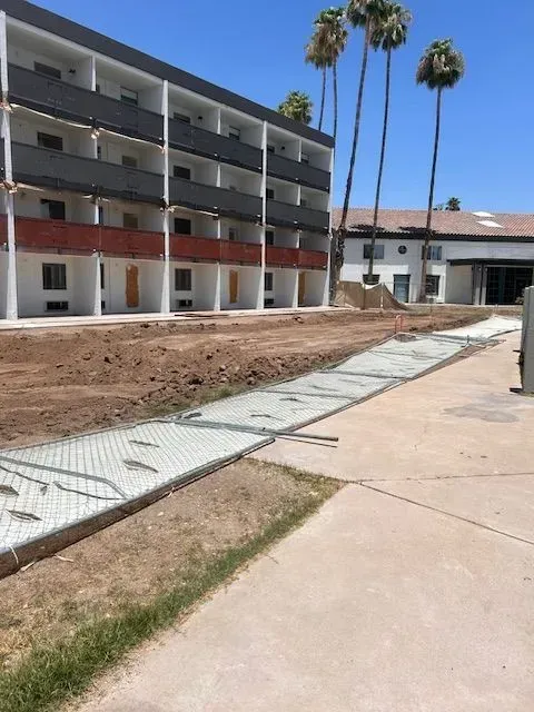 Construction site: hotel building with a chain link fence, dirt, and palm trees under a blue sky.