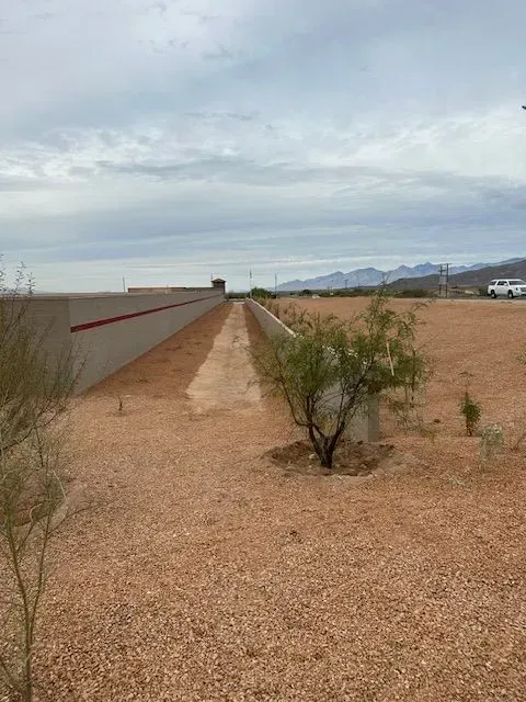 Long, dirt path between two walls, with sparse vegetation and distant mountains under a cloudy sky.