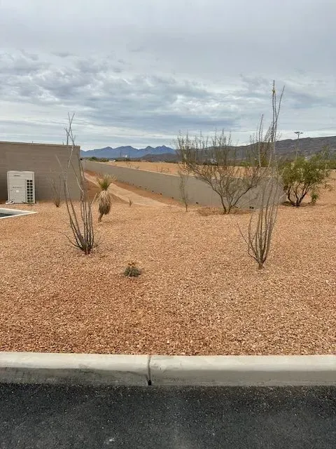 Desert landscape with tan gravel, sparse plants, a wall, and distant mountains under a cloudy sky.