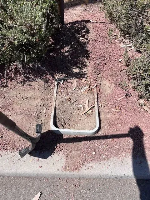 Shovel next to a rectangular irrigation valve box in the dirt next to a sidewalk with bushes.