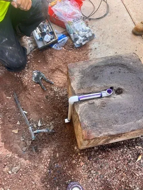 Plumber working on a water line in a dirt trench with tools and supplies nearby.