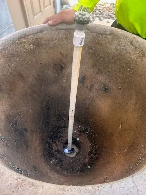 Inside a large, dark concrete pot, a white pipe with a nozzle is centered. A person's hand is visible at the edge.