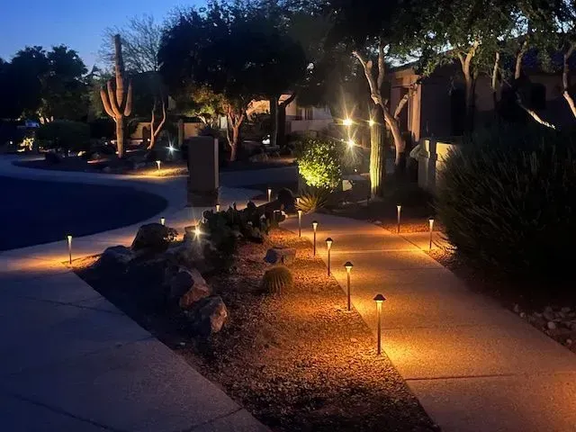 Pathway lined with small, warm-toned lights, illuminating a desert landscape at dusk.