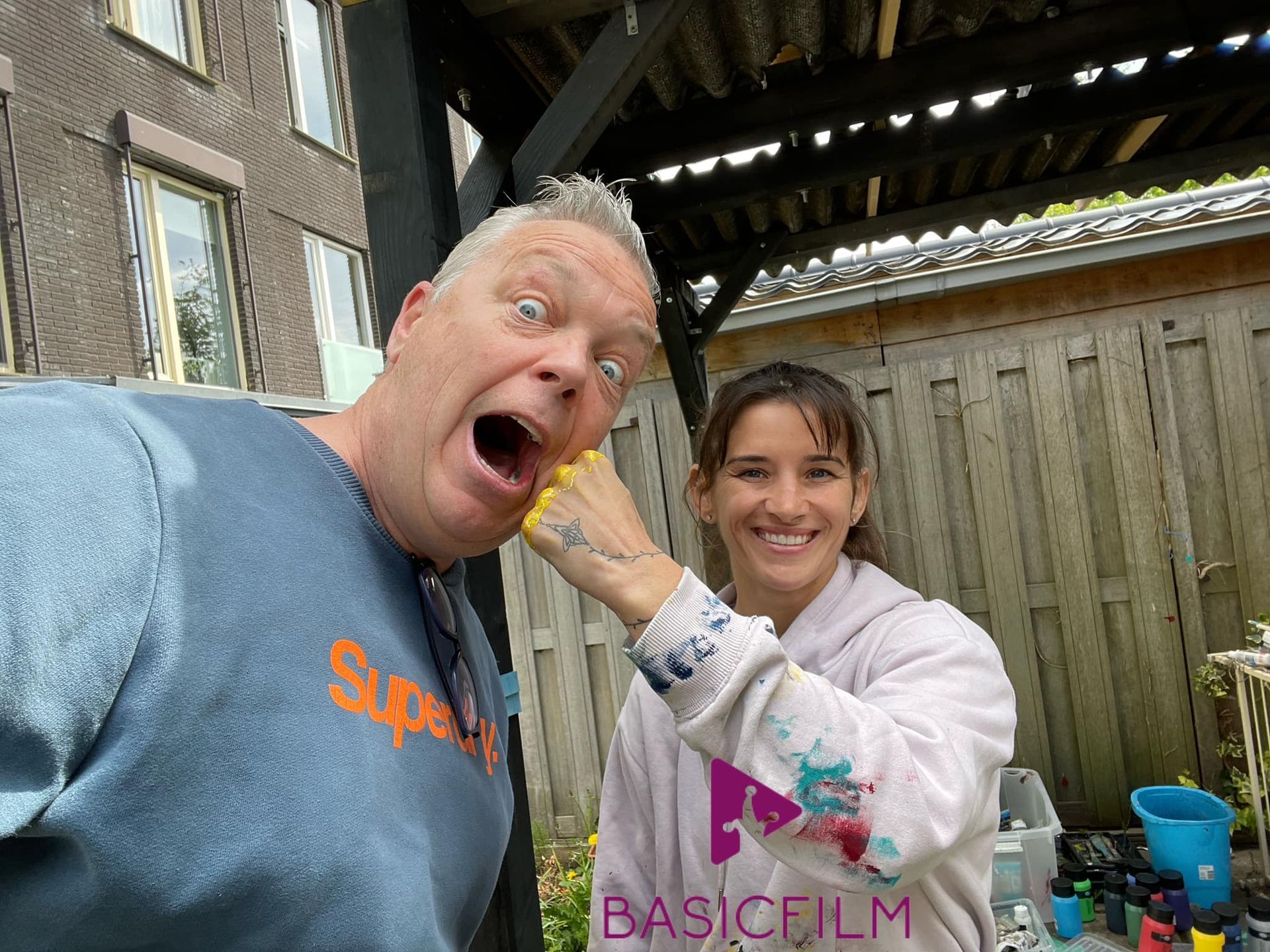 Man met overdreven gezichtsuitdrukking terwijl vrouw speels zijn gezicht aanraakt met haar geverfde hand, buitenomgeving.