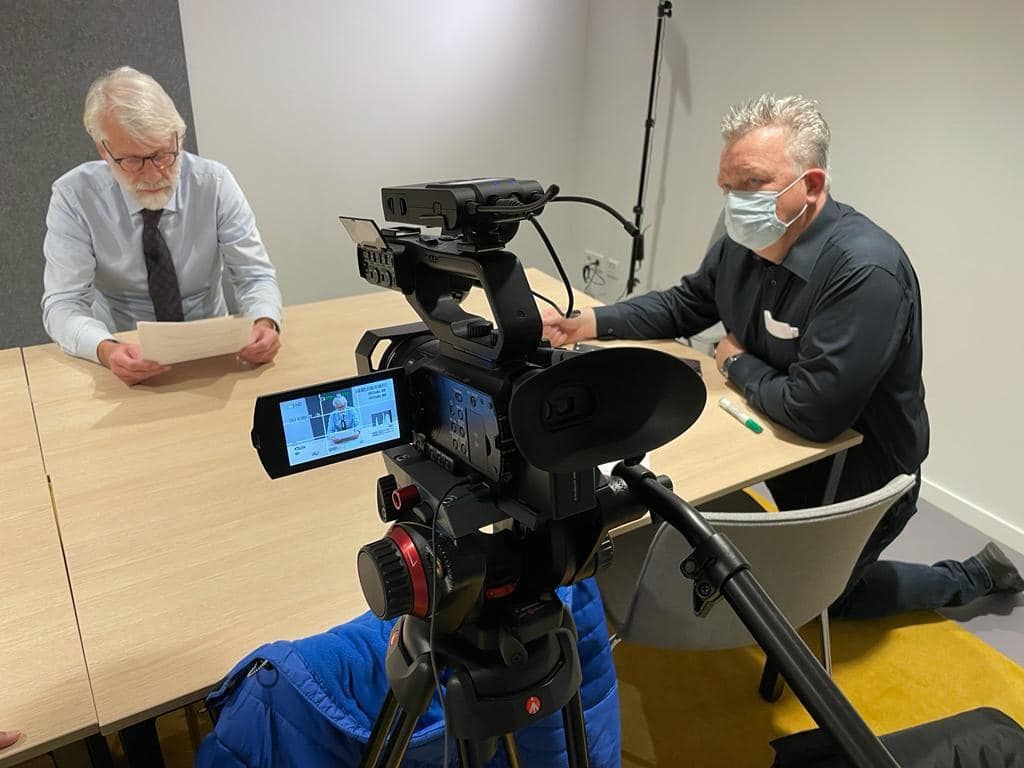 Een videocamera filmt twee mannen die aan een tafel zitten. Eén man draagt ​​een masker. De ander houdt papieren vast.