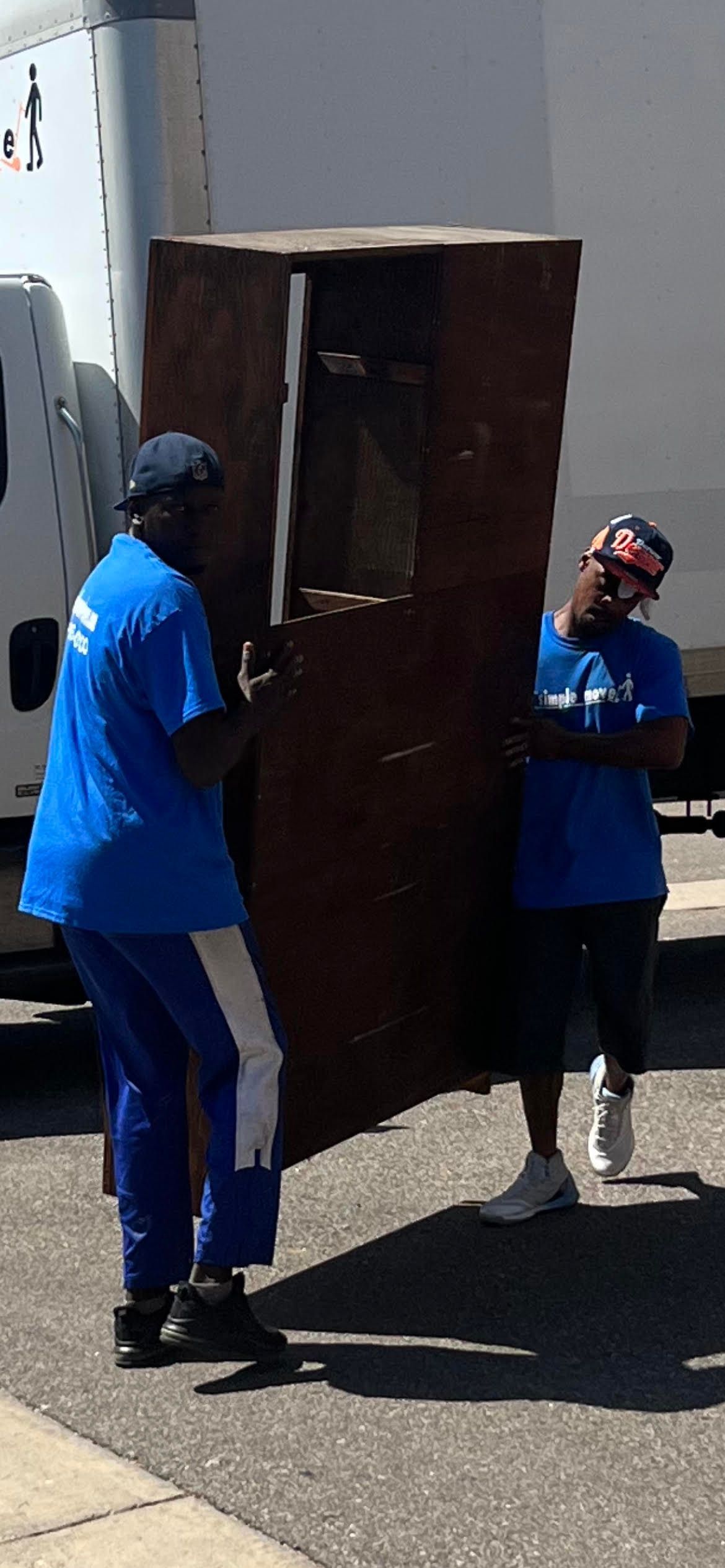 Two men in blue shirts are carrying a large wooden cabinet.