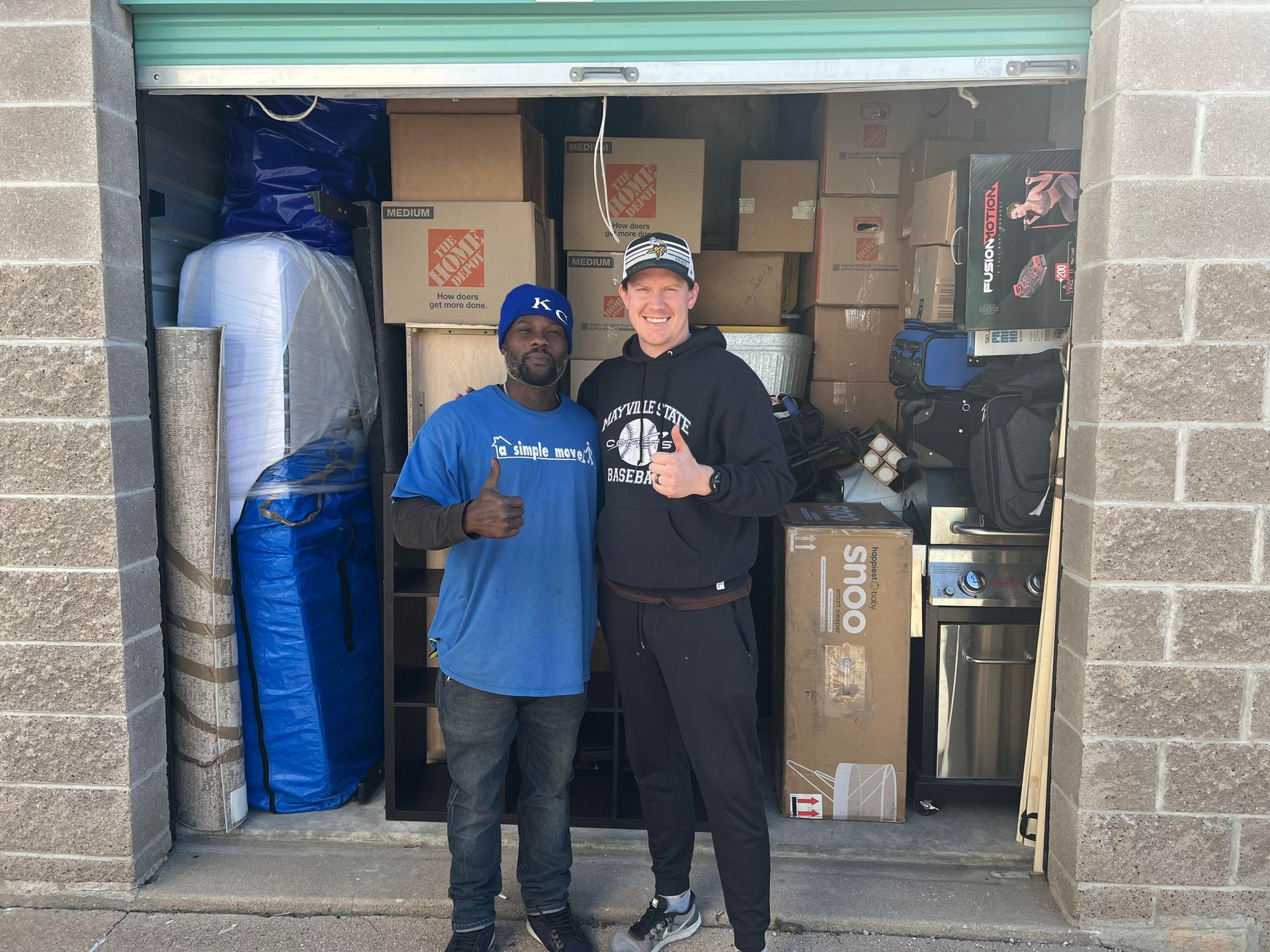 Two men are standing in a garage filled with boxes and giving a thumbs up.