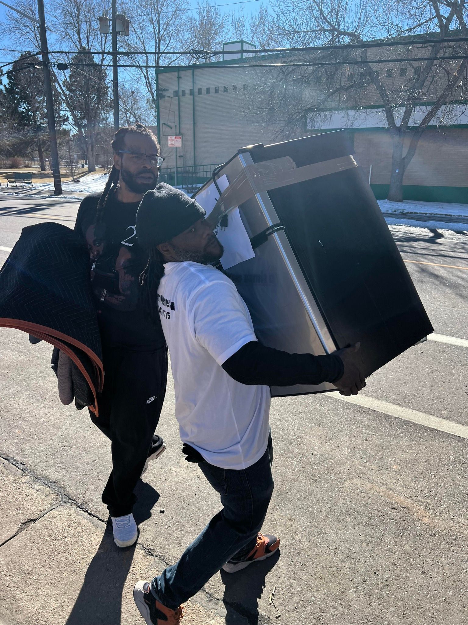 Two men are carrying a refrigerator down a street.