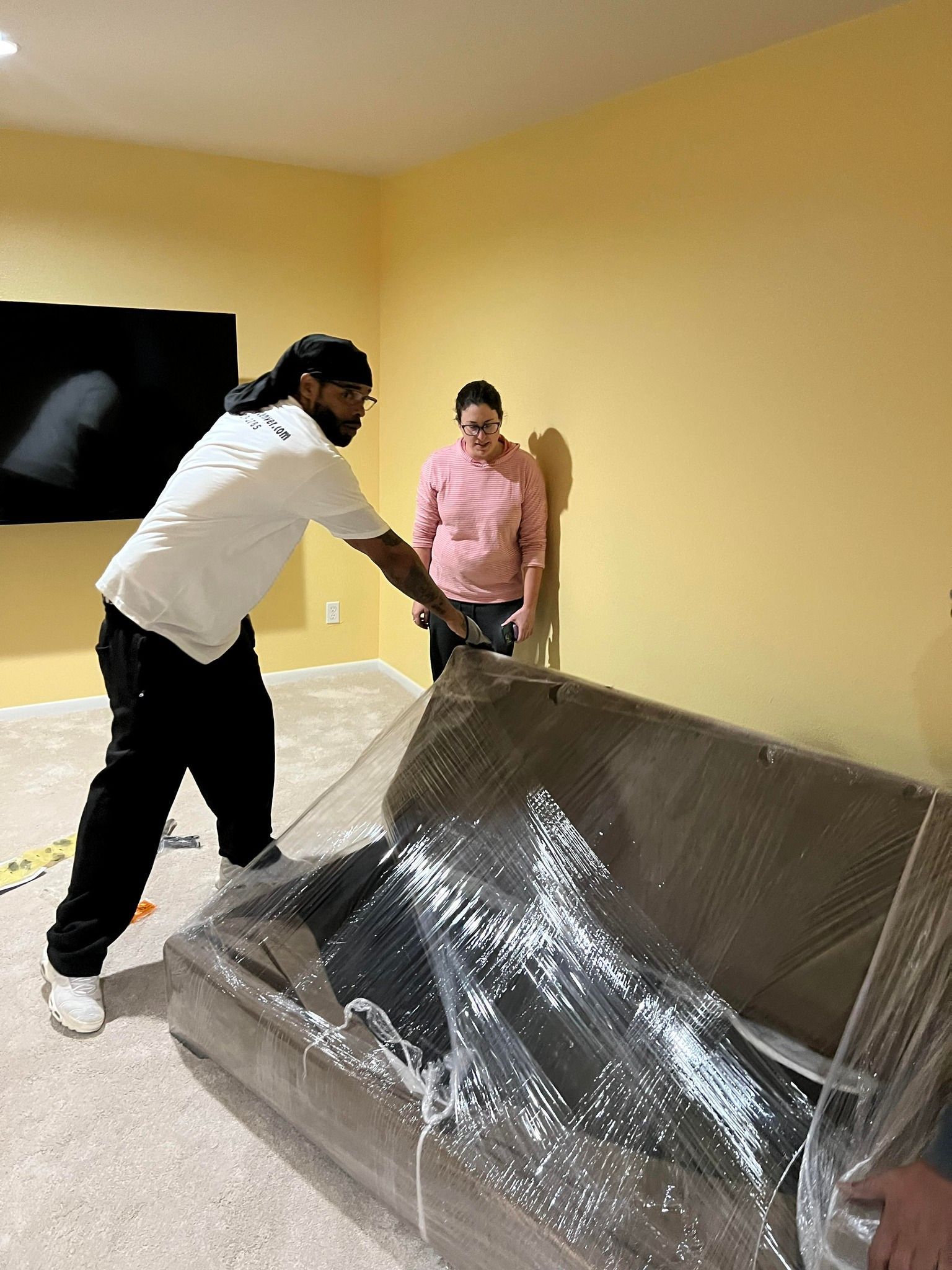 A man is wrapping a couch in plastic while a woman looks on.