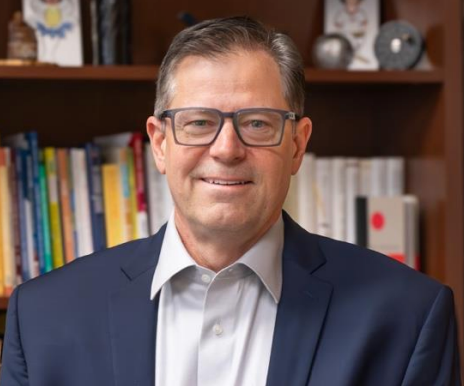 A man in a suit and glasses is sitting in front of a bookshelf.