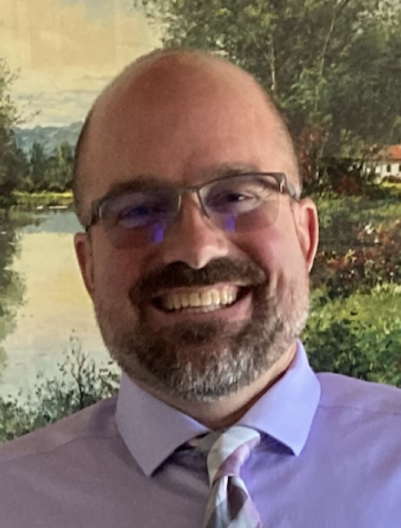 A bald man with a beard and glasses is smiling in front of a painting.