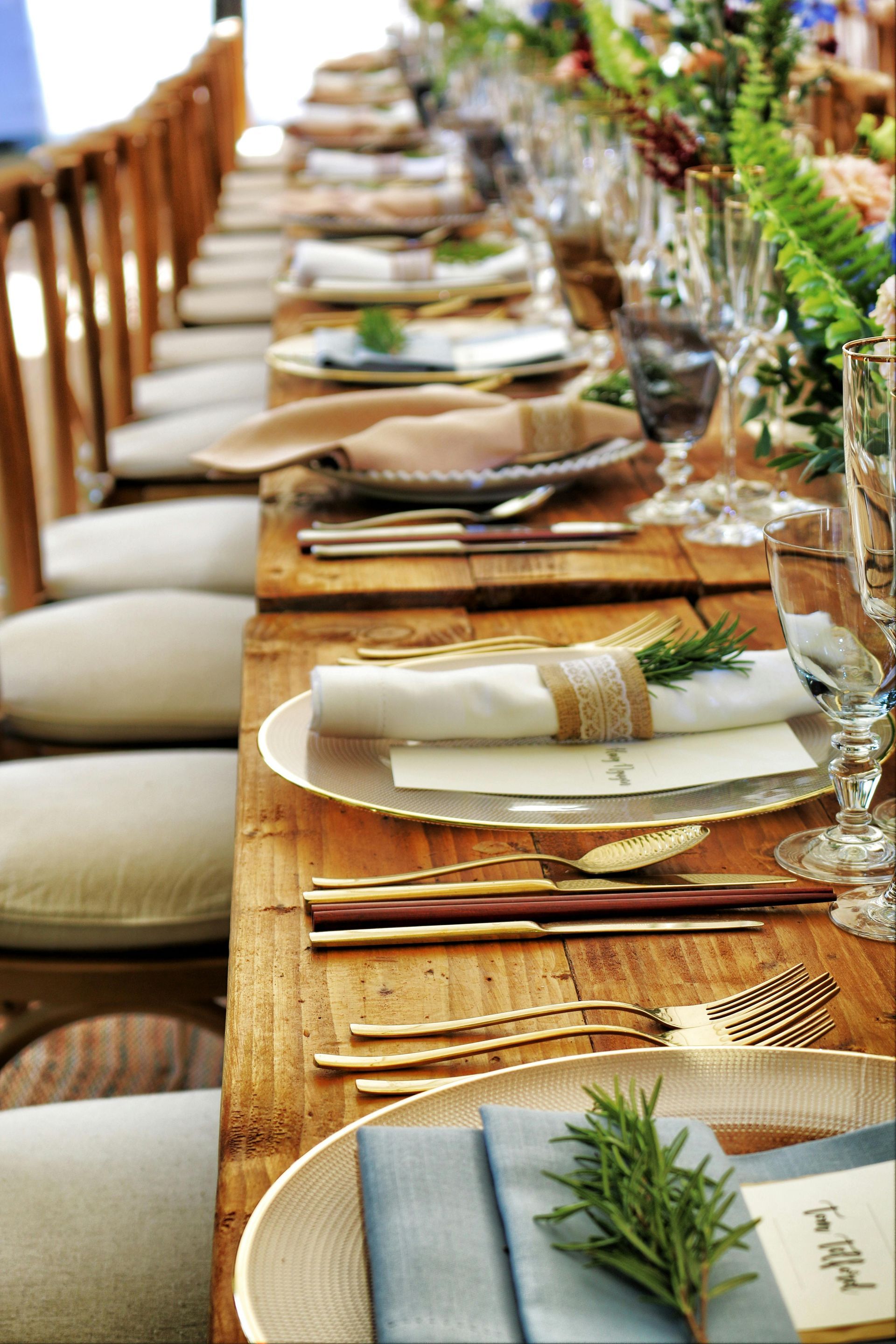 Long banquet table set with plates, napkins, glassware, and greenery centerpiece outdoors