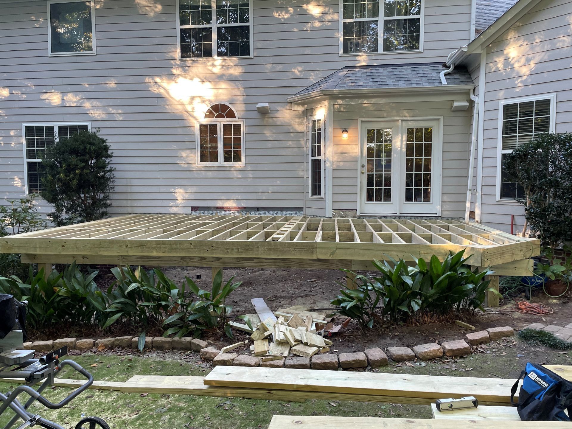 Deck frame construction in backyard, connecting to a light-colored house with multiple windows and doors.