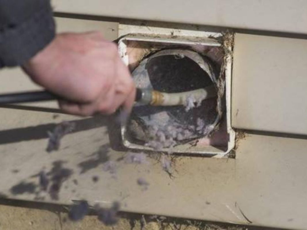 Person cleaning a dryer vent, removing lint from the exterior outlet on a beige siding.