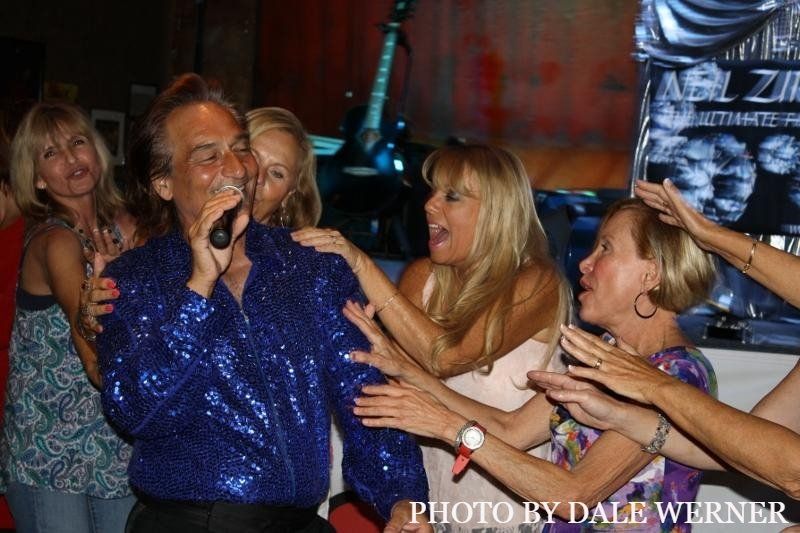 A man in a blue sequined shirt is singing into a microphone surrounded by women