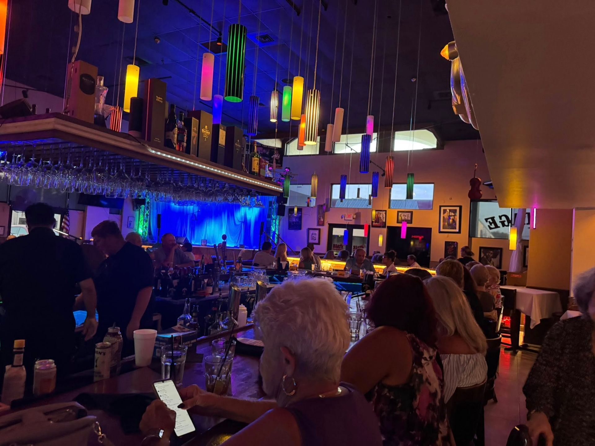 Bar interior, patrons at counter, colorful hanging lights, blue backlit stage.