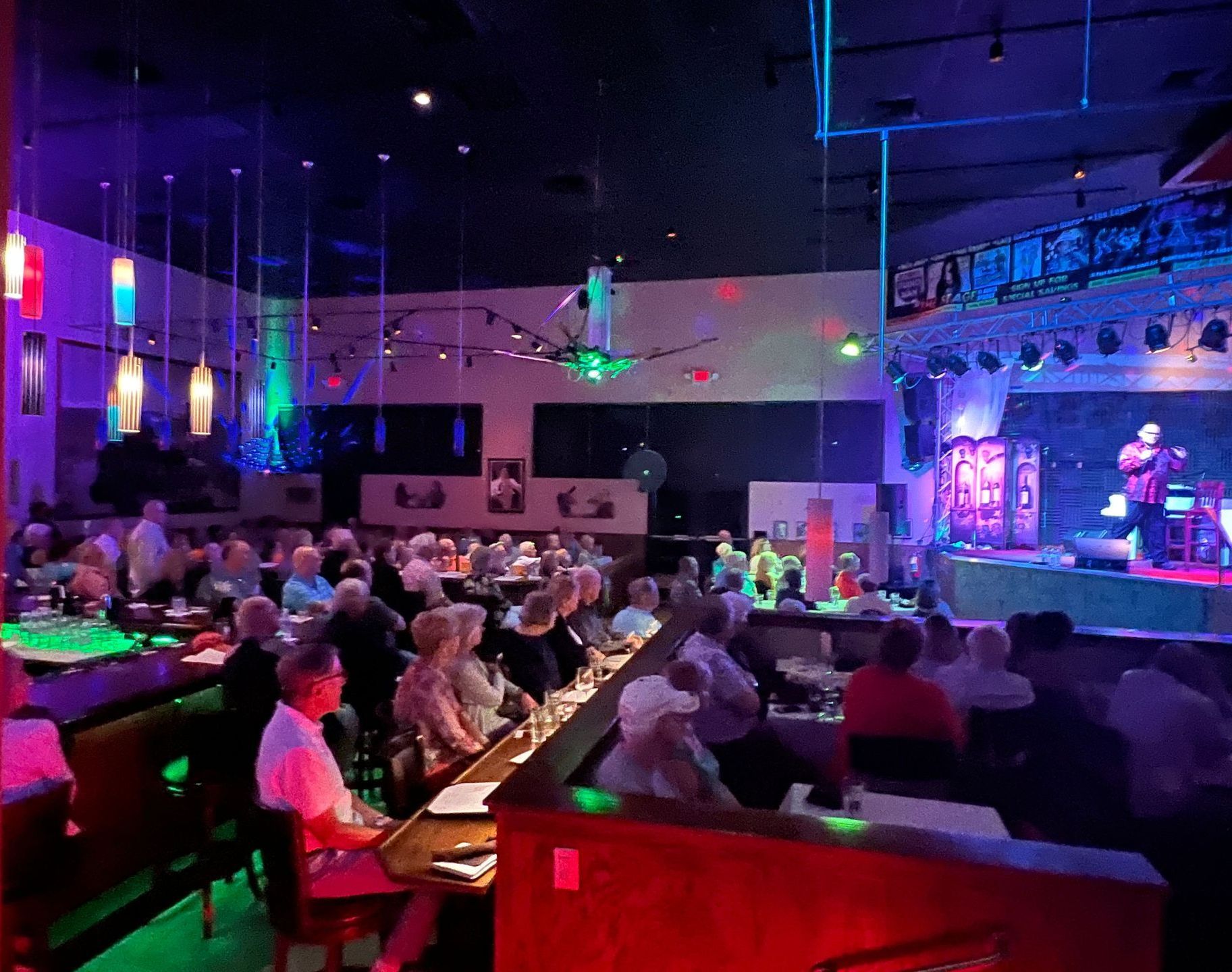 Audience watches a performer onstage in a dimly lit venue with colorful stage and overhead lighting.