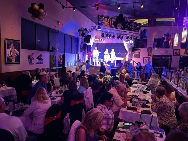 A large group of people are sitting at tables in a restaurant watching a band on stage.