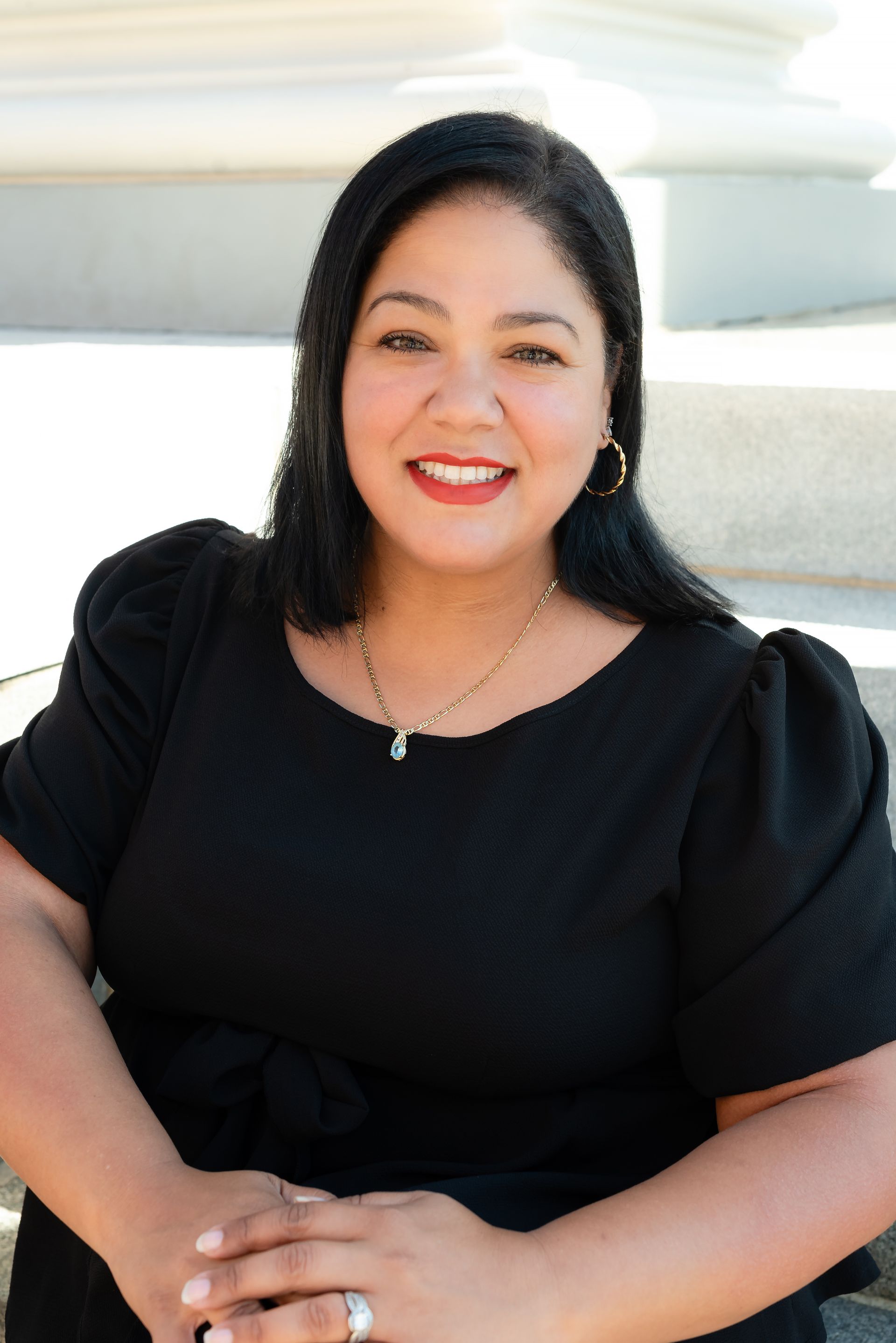 A woman in a black shirt is sitting down with her hands folded and smiling.