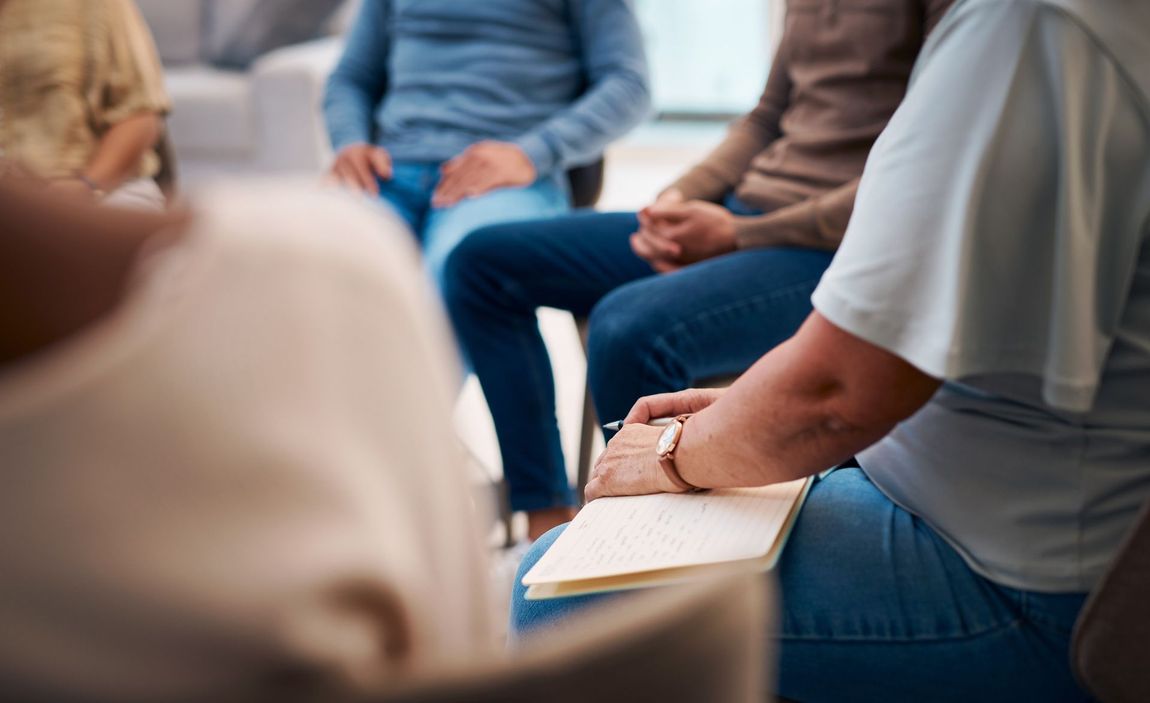 Persone sedute in cerchio durante una seduta di terapia di gruppo, alcune con in mano dei quaderni.