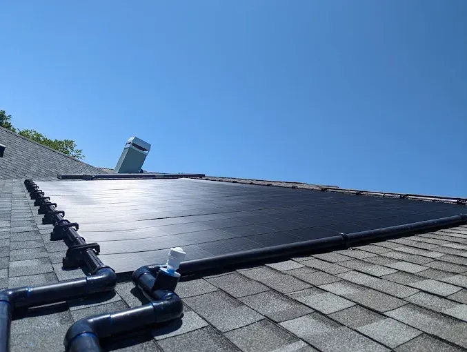 Solar panels on a shingled roof with dark blue piping against a clear, blue sky.