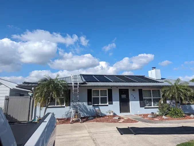 A house with solar panels on its roof, a ladder, and a partly cloudy sky.