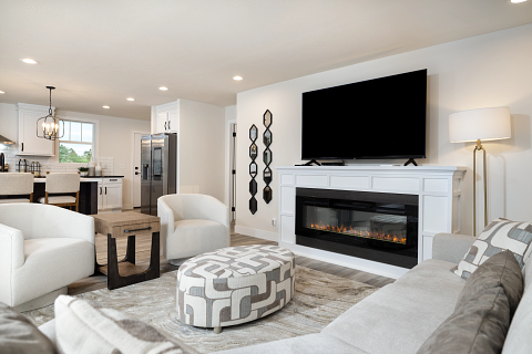 Living room with white furniture, a fireplace, and a large TV. Kitchen visible in the background.