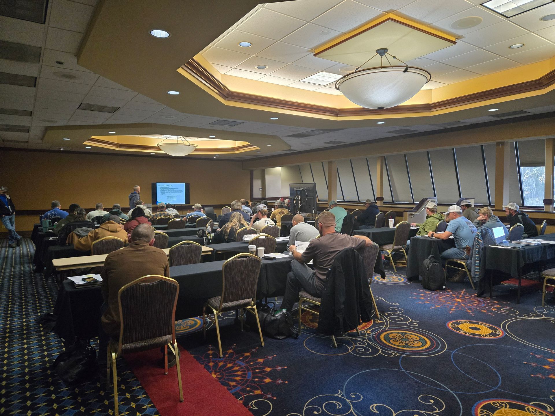 Conference room with attendees seated at tables, a speaker at the front, and a presentation on screen.