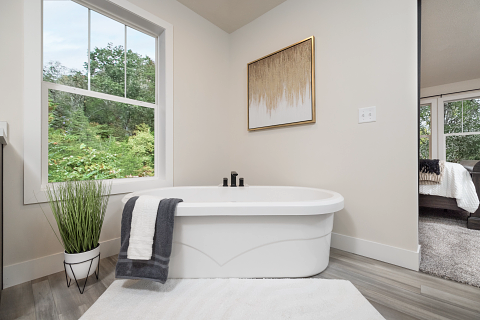 White oval bathtub in a bright bathroom with window, greenery, art, and a large mirror reflecting a bedroom.