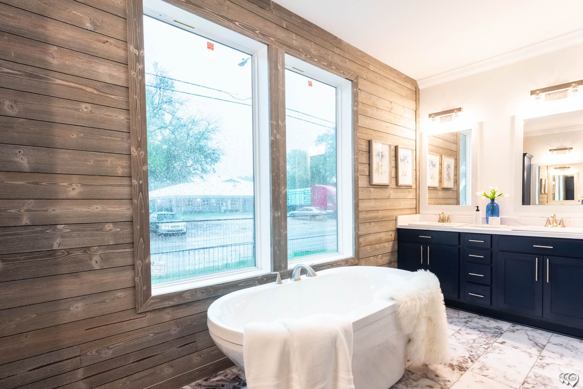 Bathroom with a soaking tub in front of a window, rustic wood wall, and navy blue vanity.