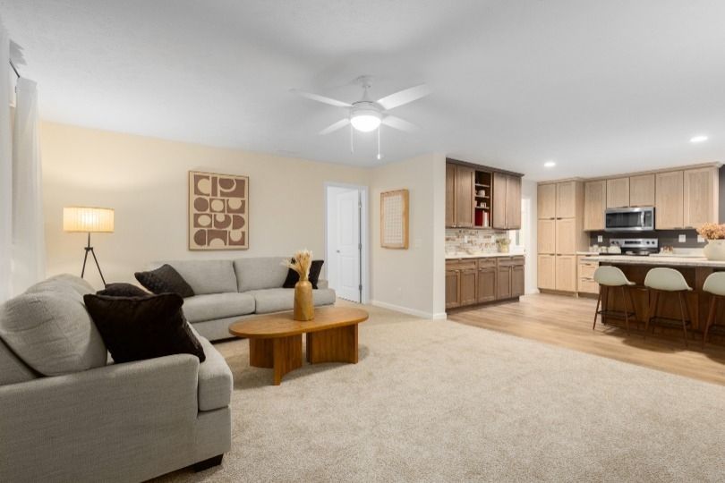 Open-concept living room and kitchen; light beige walls, gray sofa, wooden table, and kitchen cabinets.