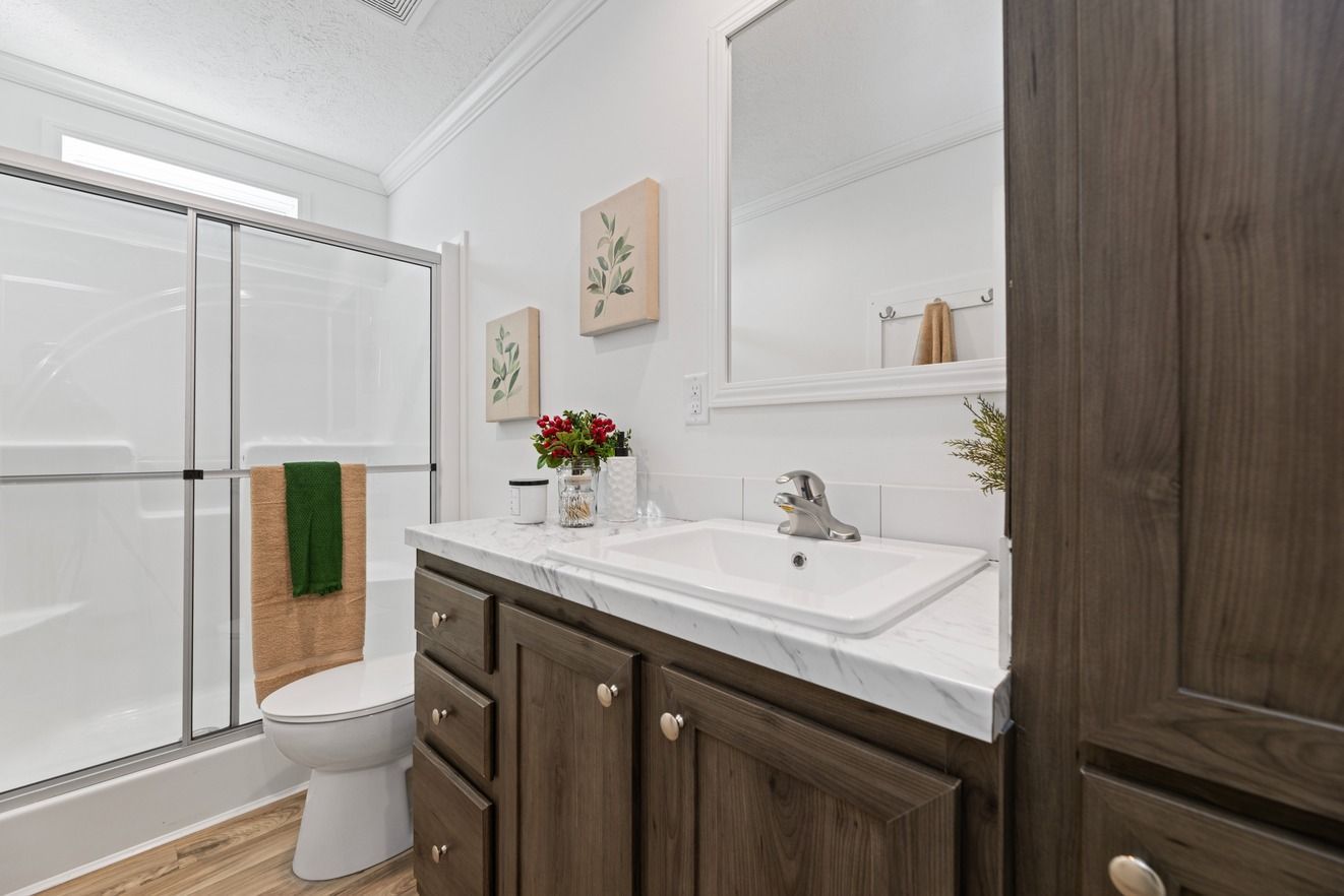 Bathroom with white walls, shower, vanity, and brown cabinets. Two framed pictures and greenery on the countertop.