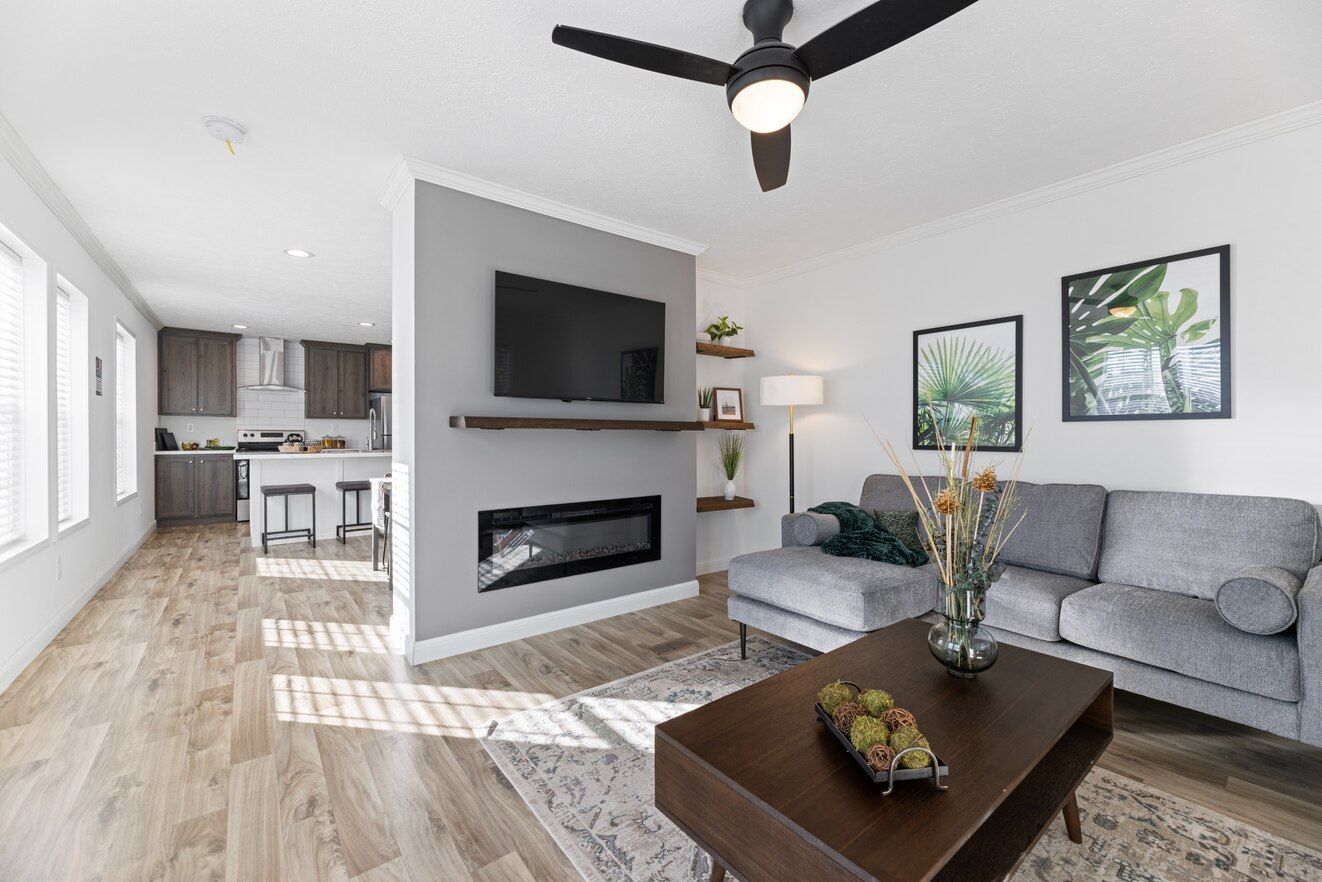 Modern living room with fireplace, TV, gray couch, and wooden floor. Kitchen visible in the background.
