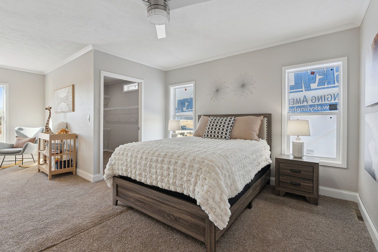Bedroom with bed, nightstand, windows, and light-colored carpet. A desk and a doorway to a walk-in closet are also visible.