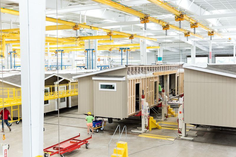 Factory with modular homes being assembled; workers, cranes, and building materials are present.
