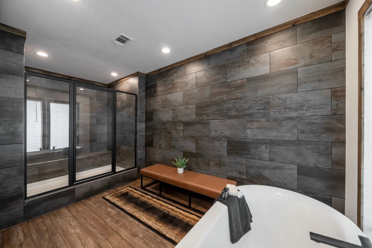 Modern bathroom with dark tile walls, glass shower, and freestanding tub. A bench and rug sit in front of the wall.