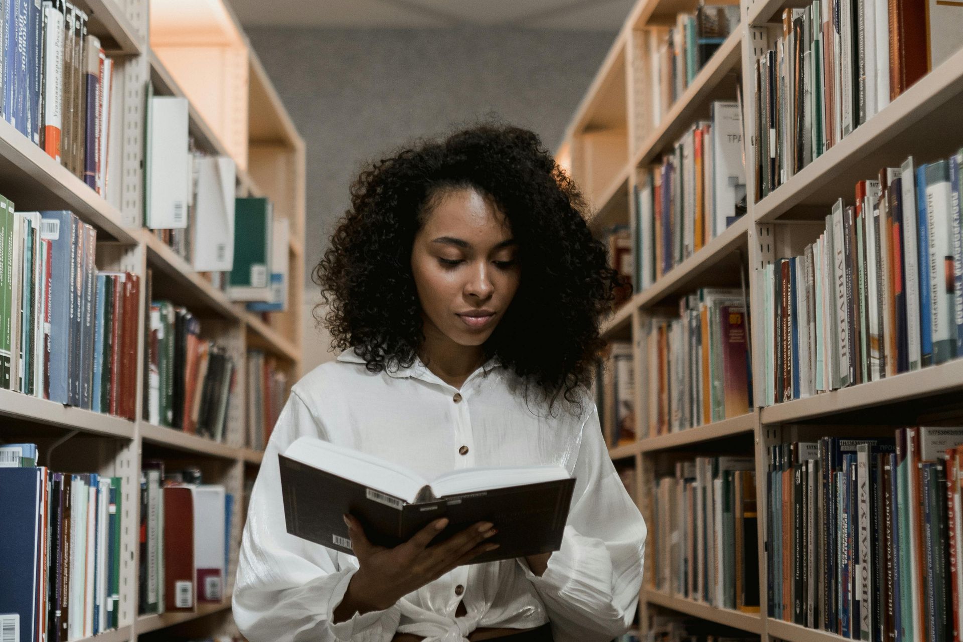 Woman reading a book in a library, surrounded by bookshelves.