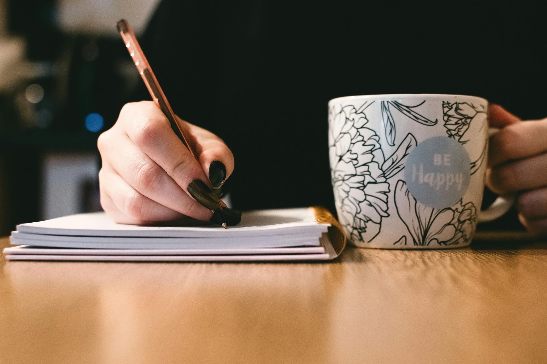 Person's hand with black nails writing in a notebook next to a floral mug labeled "BE HAPPY."
