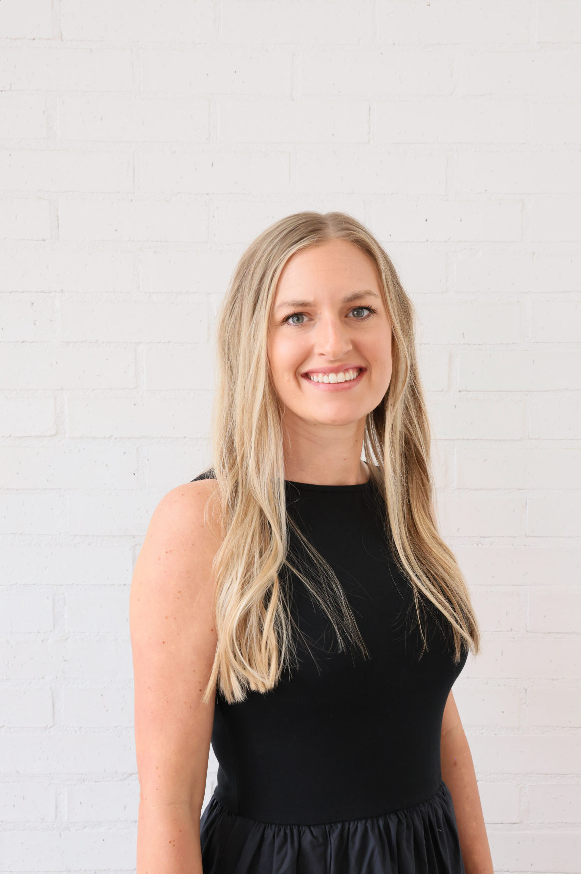 A woman in a black dress is smiling and standing in front of a white brick wall.