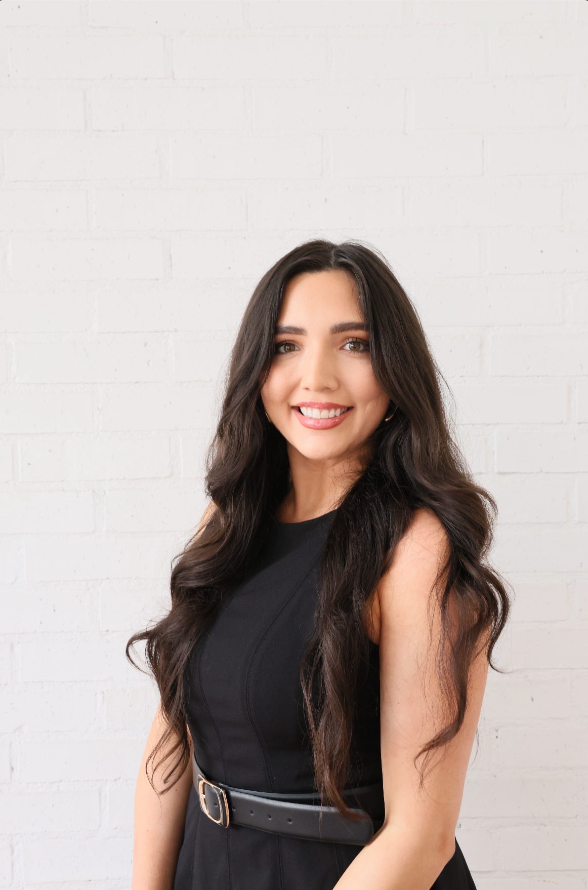 A woman in a black dress is smiling and standing in front of a white wall.