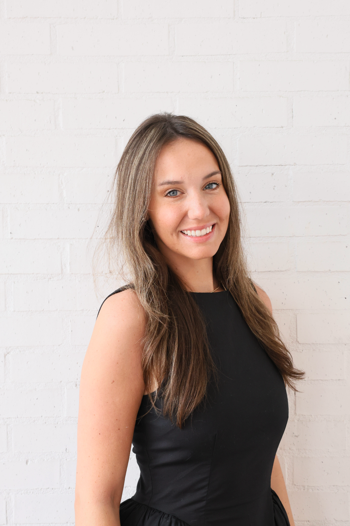 A woman in a black dress is smiling and standing in front of a white brick wall.