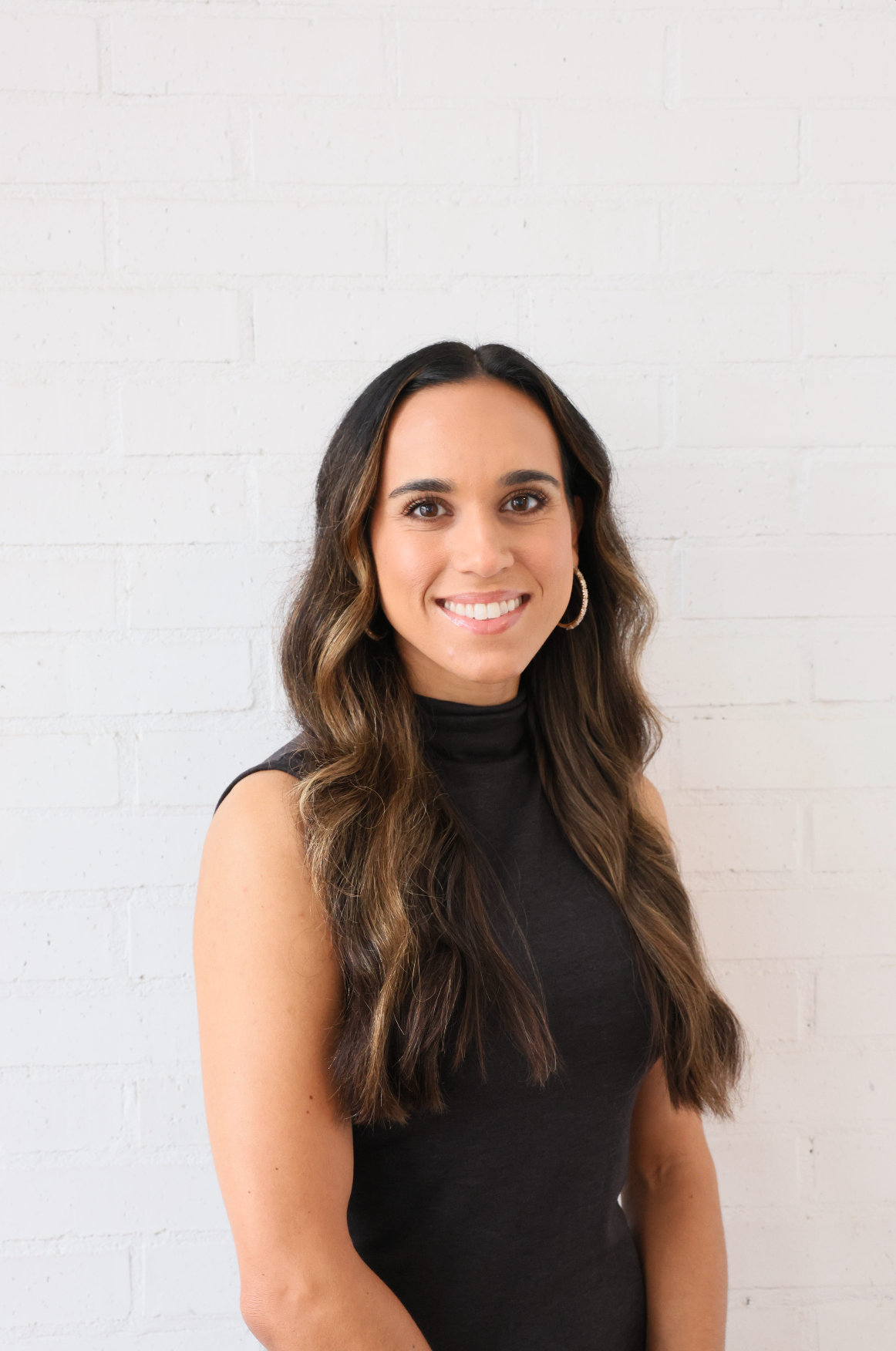 A woman in a black dress is smiling and standing in front of a white brick wall.