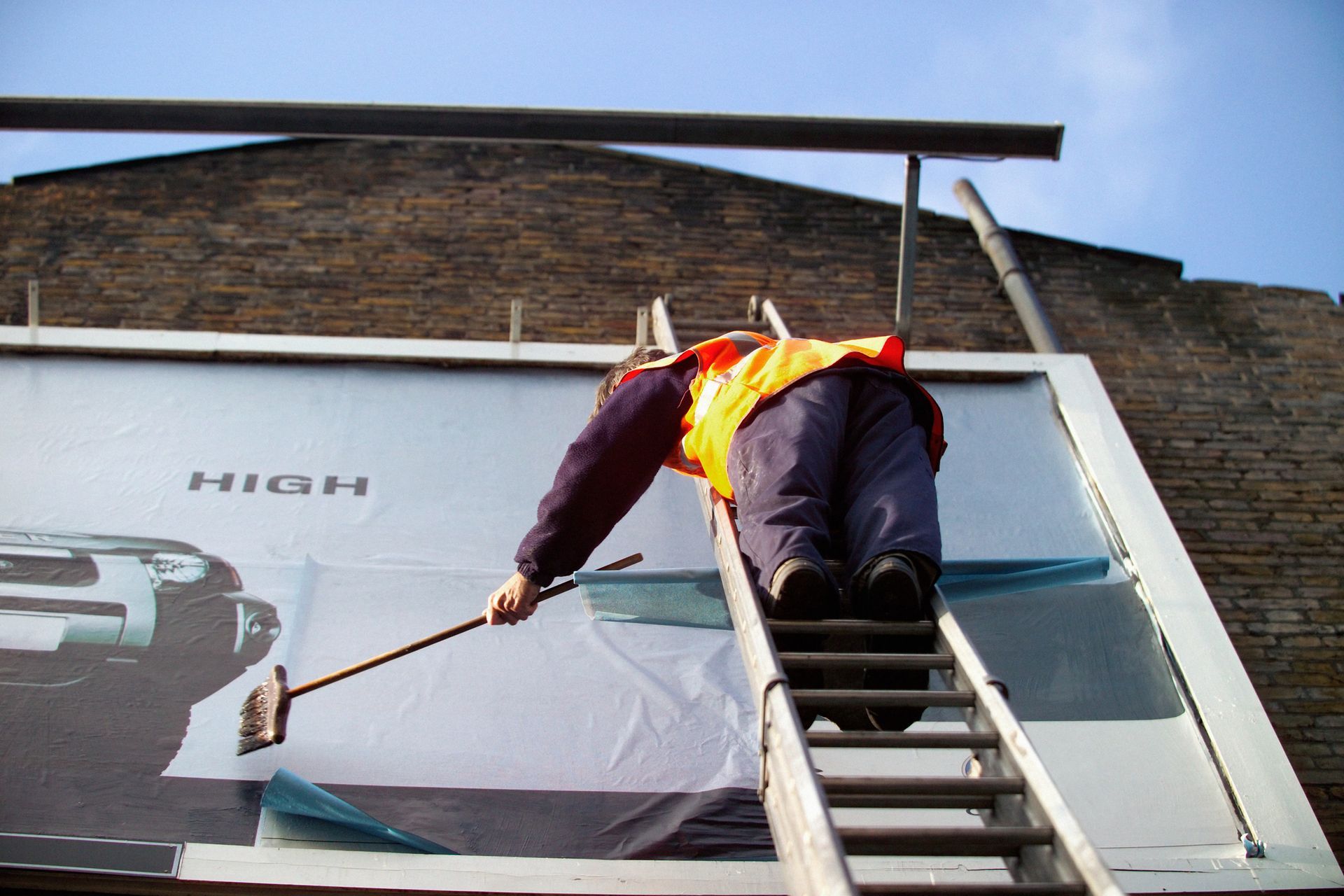 A sign installer on a ladder is working on a commercial sign by the side of a building.