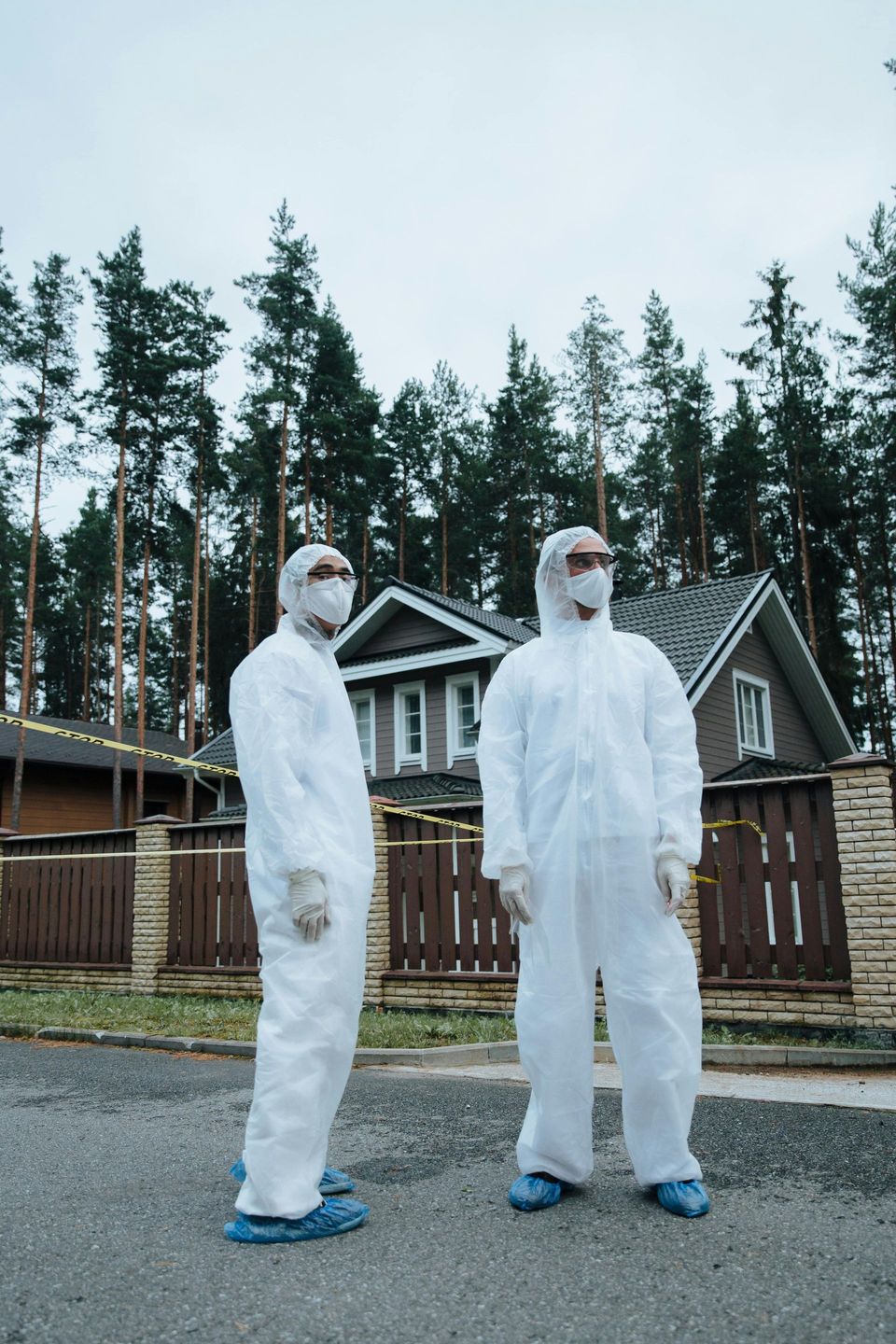 Two people in white hazmat suits stand in front of a house, yellow tape surrounds the property.