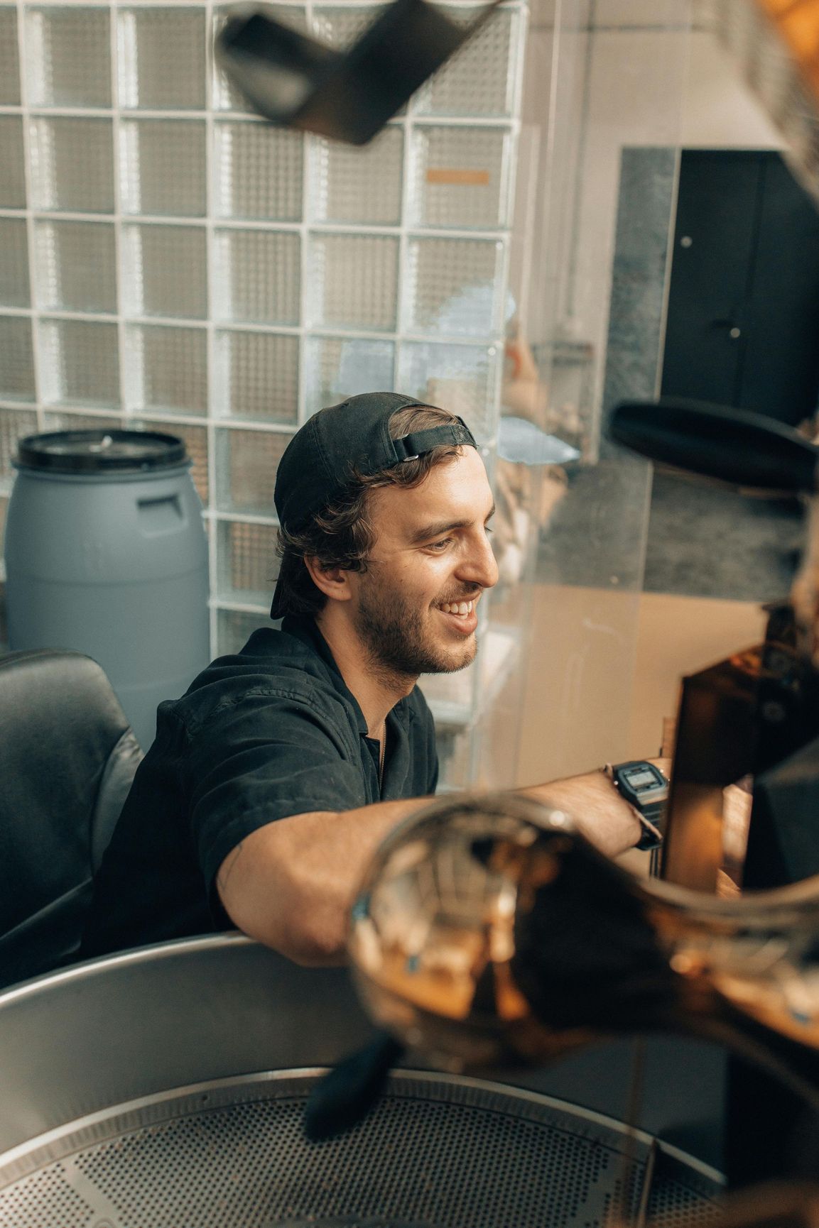 Man in black shirt and cap smiles, working with metal machinery in a warehouse-like setting.