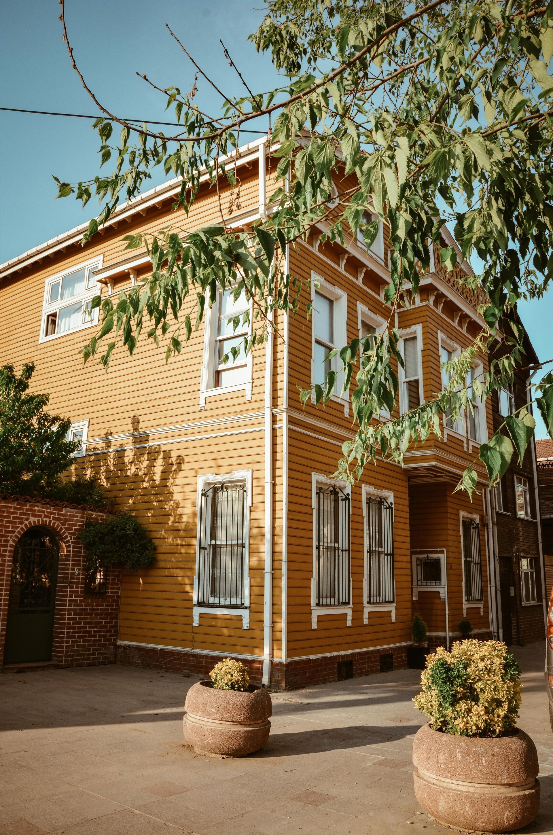 A three-story wooden house, painted yellow, with white-framed windows; leafy trees surround it.