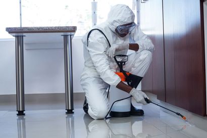 Person in protective suit spraying insecticide near a cabinet and table.